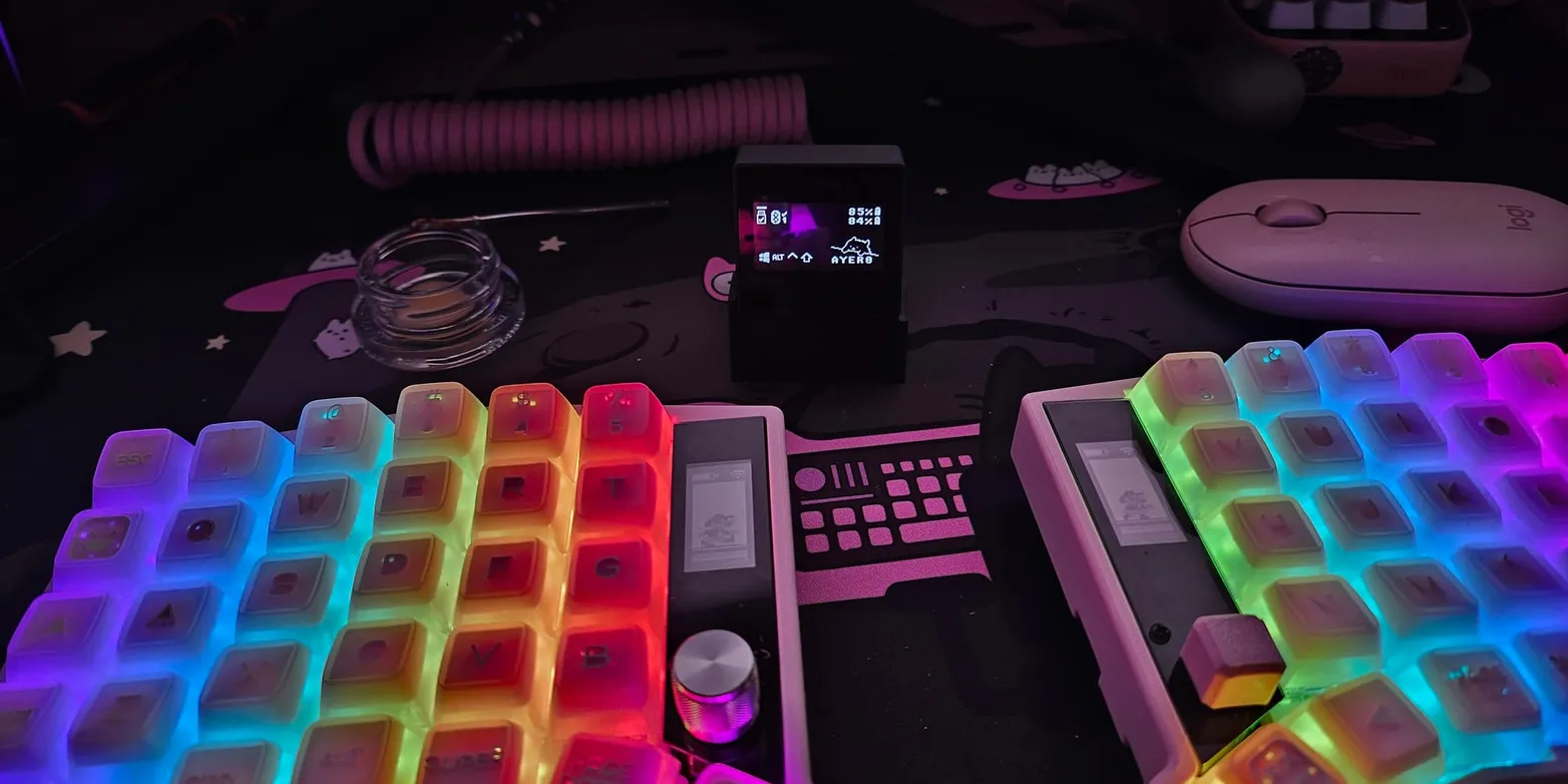 A wireless Sofle split keyboard with rainbow RGB backlighting and a rotary knob, next to a small dongle screen showing battery levels for both halves