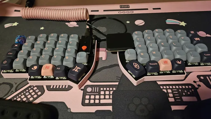 A Lily58 split keyboard in a pink case with grey and navy keycaps, a red trackpoint on the left half and a square touchpad module between the halves