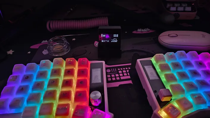 A wireless Sofle split keyboard with rainbow RGB backlighting and a rotary knob, next to a small dongle screen showing battery levels for both halves