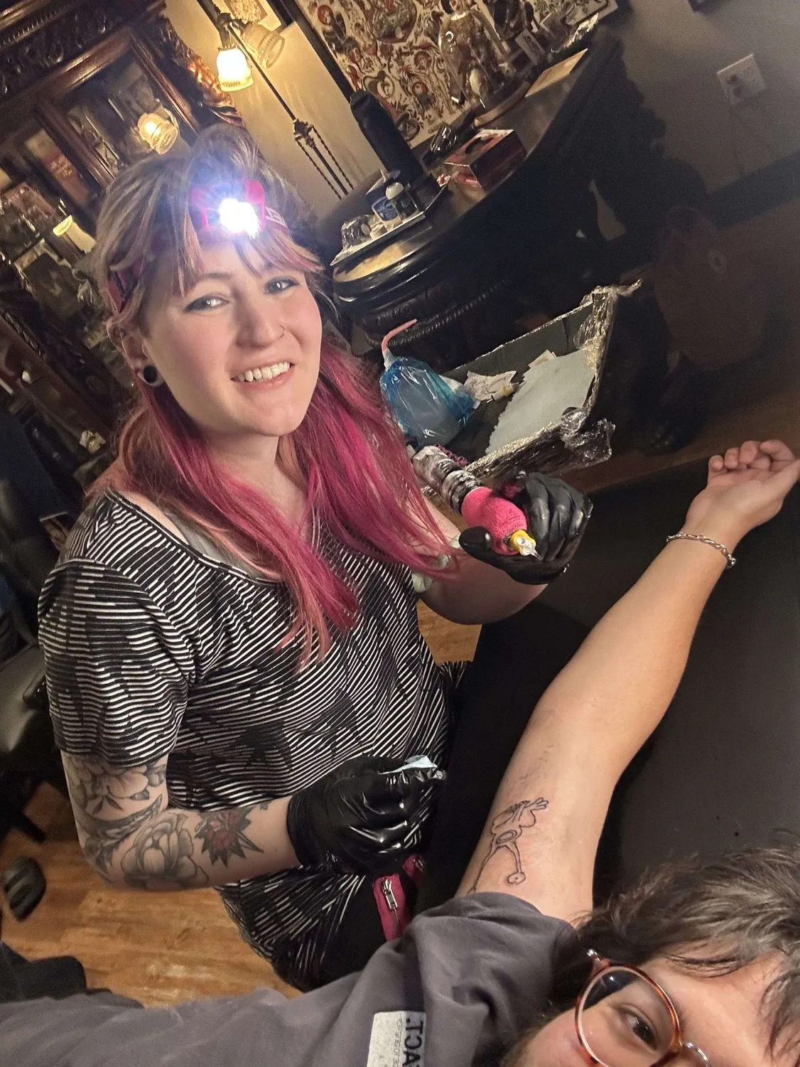 Allison in a tattoo studio wearing a headlamp and holding a pink tattoo machine, next to a client's forearm with a fresh line-work frog tattoo