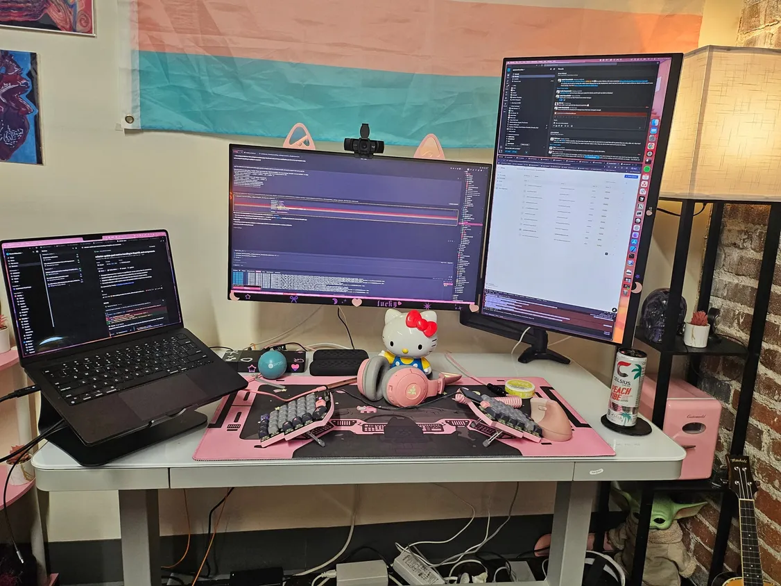 Allison's desk with a laptop and two monitors above a pink split keyboard on a pink desk mat, next to a Hello Kitty figure
